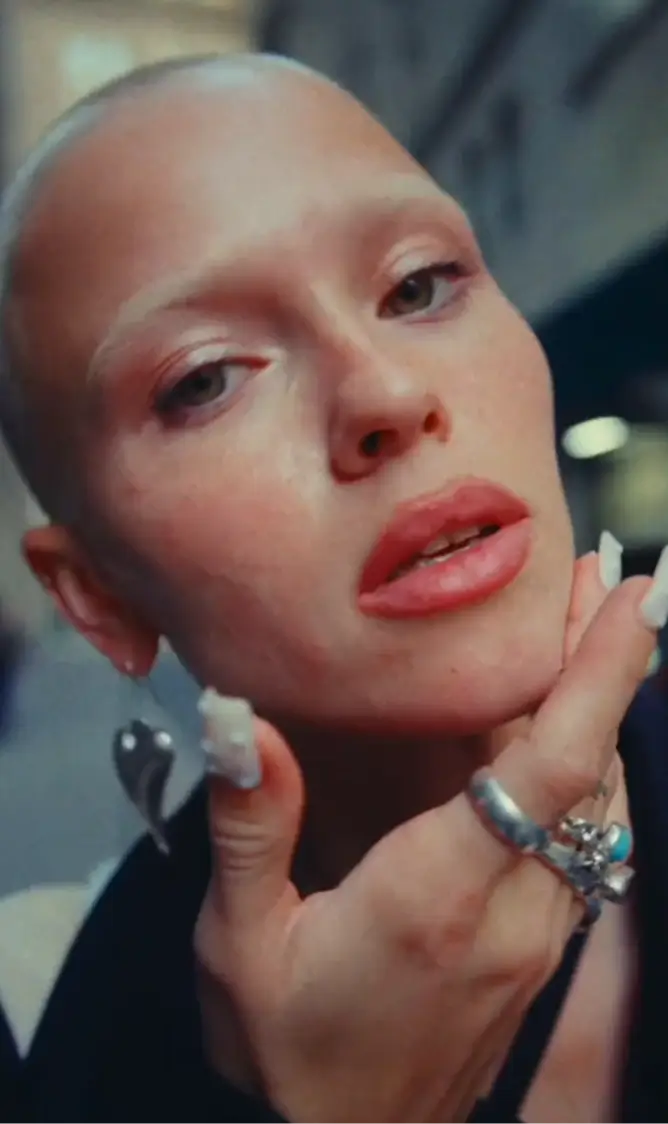 Close-up of a person with a shaved head, light makeup, and silver earrings, touching their chin with rings on their fingers.