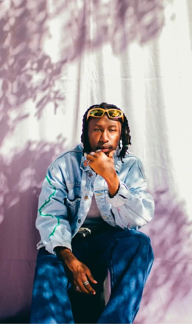 Man with braided hair wearing yellow sunglasses on head, a light denim jacket, and blue jeans sitting thoughtfully against a white backdrop with purple shadow patterns.